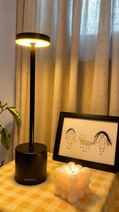 Black Egret Lamp on a gingham tablecloth beside a framed child's drawing and a lit candle