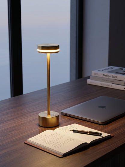Gold Alba Lamp on a wooden desk beside a laptop and an open notebook by a window