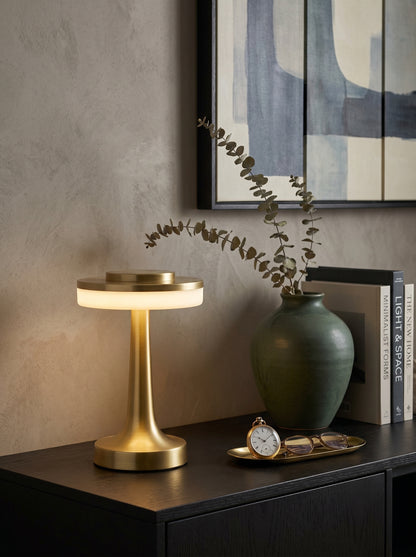 Gold Kestrel Lamp, a brushed gold lamp with a glowing ring diffuser, on a black console with a vase