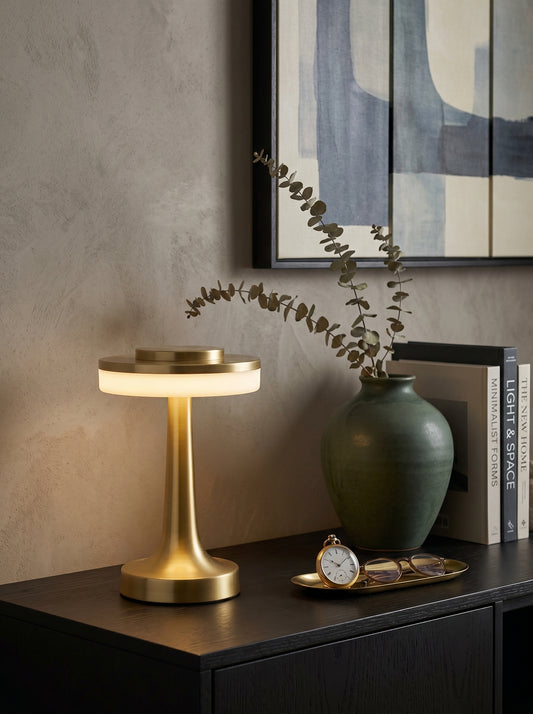 Gold Kestrel Lamp, a brushed gold lamp with a glowing ring diffuser, on a black console with a vase