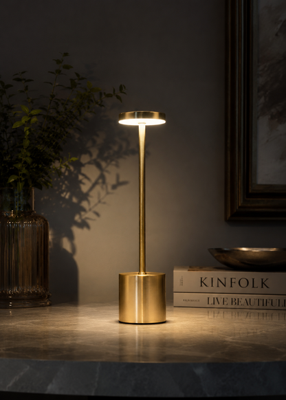 Gold Egret Lamp, a slim brushed gold cordless lamp with a glowing disc head, on a dark marble table