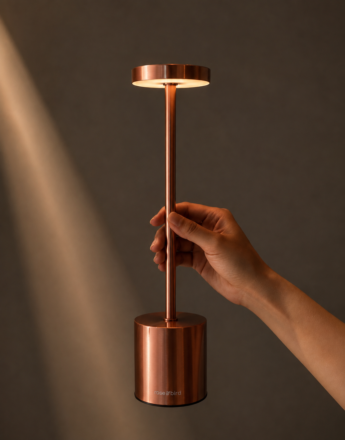 Hand holding the Rose Gold Egret Lamp, a slim cordless table lamp with a glowing disc head