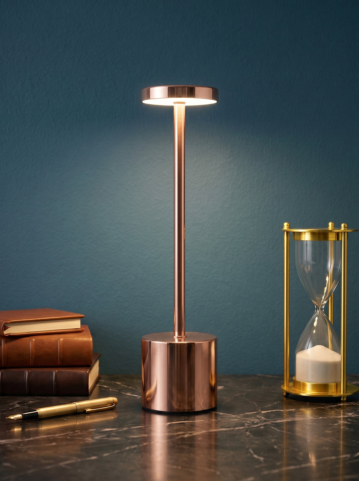 Rose Gold Egret Lamp on a dark marble desk against a teal wall with leather books and a brass hourglass