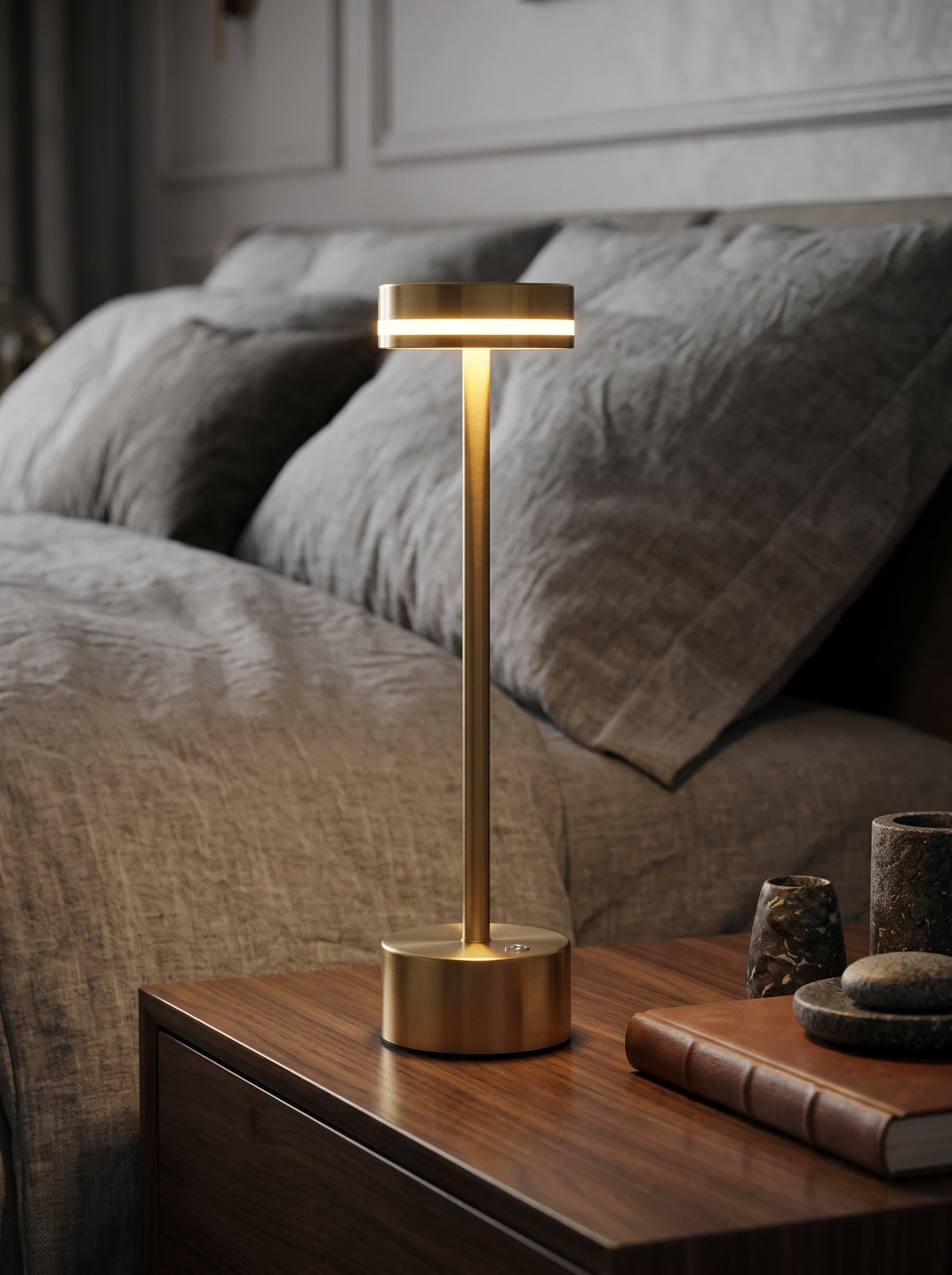 Gold Alba Lamp glowing on a wooden bedside table next to grey bedding