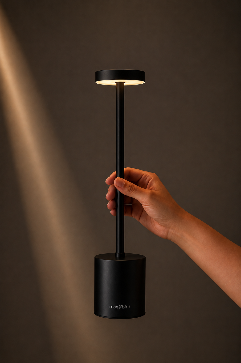 Hand holding the Black Egret Lamp, a slim matte black cordless lamp with a glowing disc head