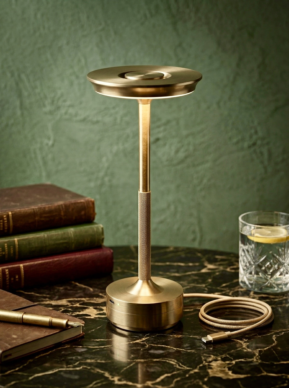 Gold Aurelia Lamp on a green marble table with leather books and a glass of water