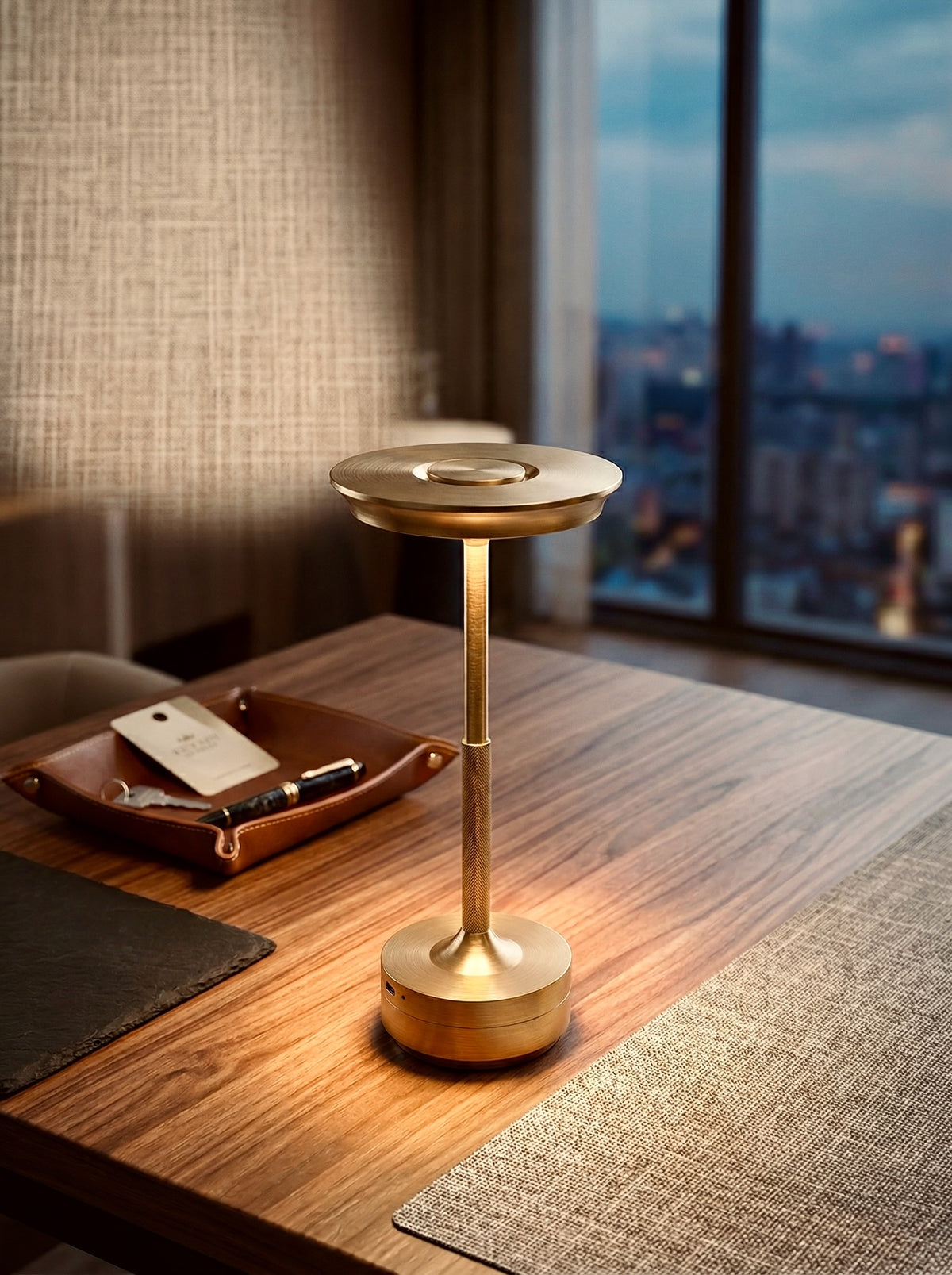 Gold Aurelia Lamp glowing on a wooden desk beside a leather tray, with a city view at dusk
