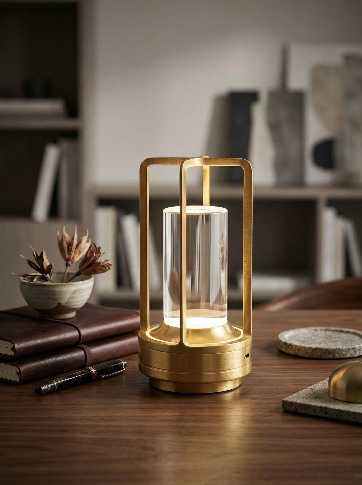 Gold Orion Lamp, a gold lantern-style lamp with a clear crystal cylinder, on a wooden desk