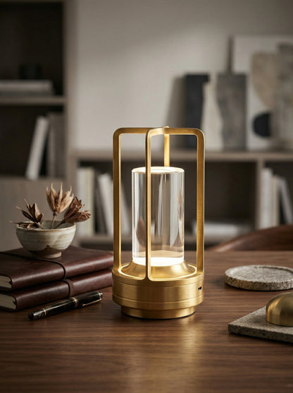 Gold Orion Lamp, a gold lantern-style lamp with a clear crystal cylinder, on a wooden desk